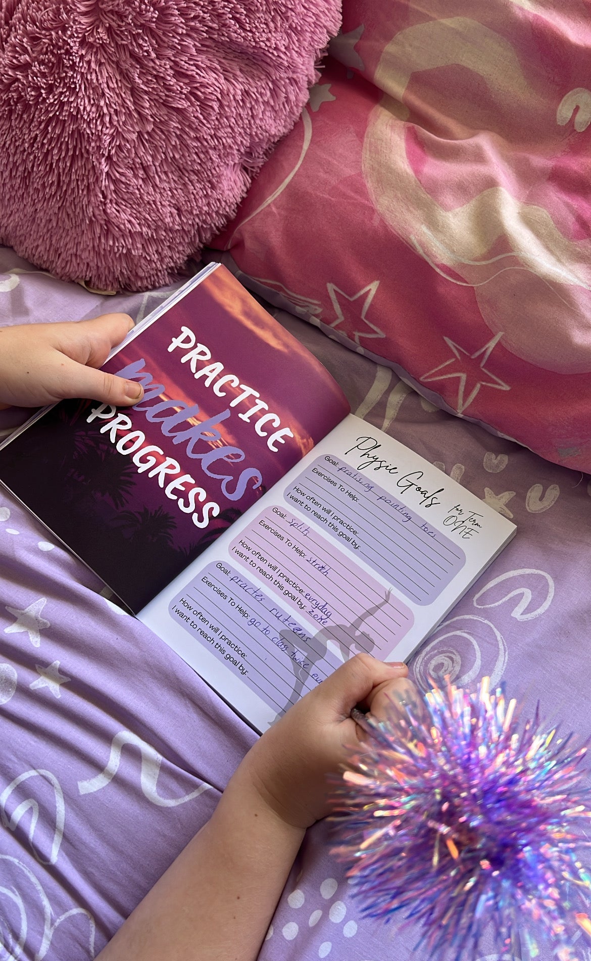 Person holding a book titled 'Practice Makes Progress' on a bed with pink pillows and a purple blanket.