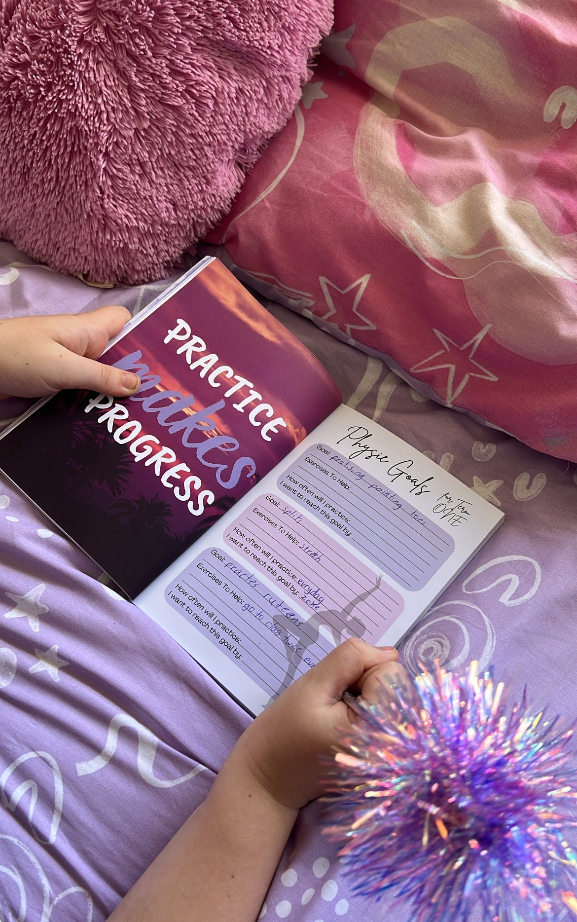 Person holding a book titled 'Practice Makes Progress' on a bed with pink pillows and a purple blanket.