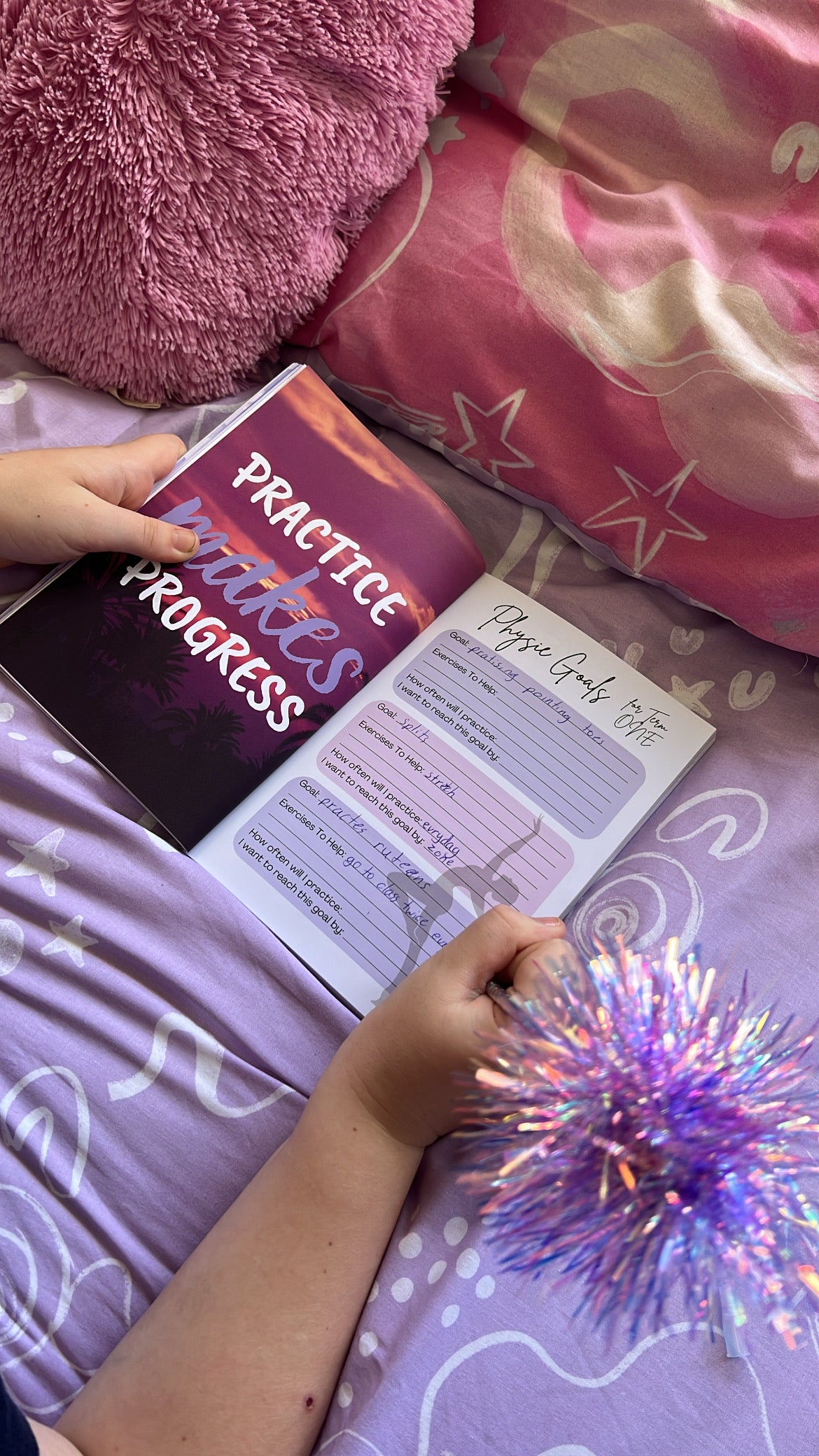 Person holding a book titled 'Practice Makes Progress' on a bed with pink pillows and a purple blanket.